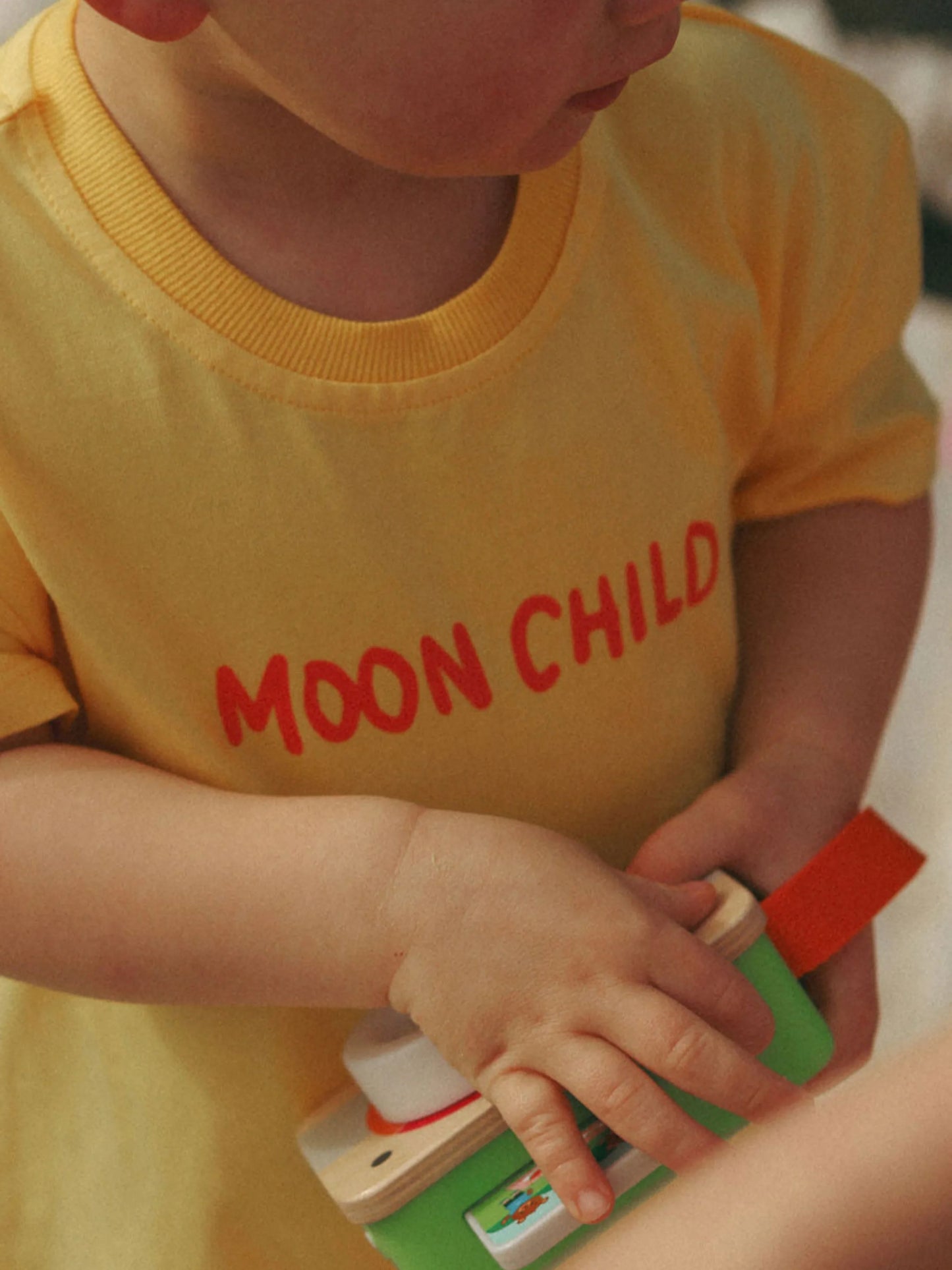 Child wearing a yellow shirt with 'MOON CHILD' text, holding a toy.